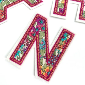 May include: A pink felt letter N with a floral pattern and gold stitching. The letter is a decorative patch or applique.