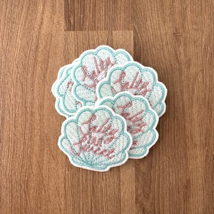 May include: A stack of six embroidered iron-on patches in the shape of a seashell with the text "Salty Hair, Don't Care" in pink lettering.