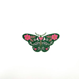 May include: A green butterfly-shaped patch with pink floral and white beaded accents. The butterfly has a detailed design with leaves and a small antenna. The patch is set against a white background.