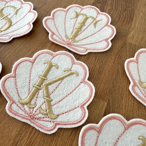 May include: Four white felt seashells with pink stitching and gold embroidered letters. The letters are K, T, S, and a letter that is not visible in the image.