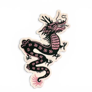 May include: An embroidered patch featuring a stylised dragon in black and pink thread. The dragon has a long, winding body with pink scale details, a detailed head with horns, and a pink tail tip, all outlined with a cream-coloured border.