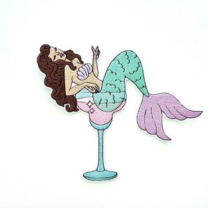 May include: A mermaid with brown hair and a green and purple tail sits in a blue and pink margarita glass. The mermaid is wearing a pink seashell bra and has a peace sign.