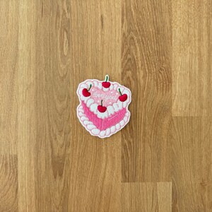 May include: Pink embroidered iron-on patch of a cake with white frosting and red cherries. The cake says "love you" in white lettering.