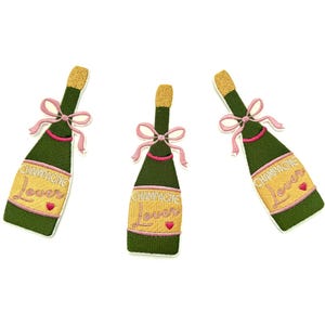 May include: Three embroidered iron-on patches shaped like champagne bottles with pink bows and the text "Champagne Lover" in pink and white lettering.