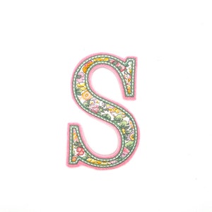 May include: A pink felt letter S with a floral pattern. The letter is surrounded by a white background.