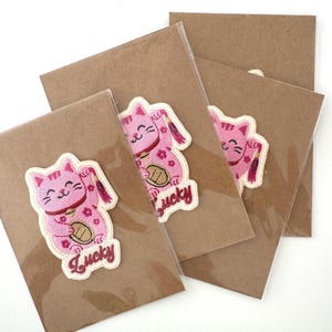 May include: Four kraft paper cards, each with a pink embroidered lucky cat patch. The patches feature a smiling cat with a raised paw, holding a gold coin, and the word "Lucky".