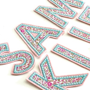 May include: A set of felt letters in pink with a floral pattern and a light blue trim. The letters spell out the name "SAIK"