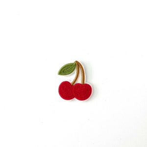 May include: A red cherry with a green leaf and a gold stem. The cherry is embroidered on a white fabric.