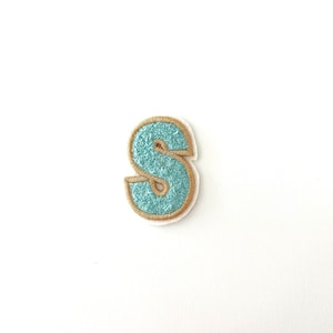 May include: A light blue felt letter S with a gold outline. The letter is embroidered and has a slightly fuzzy texture.