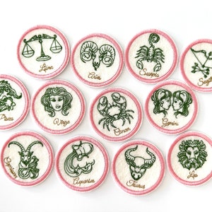 May include: Twelve embroidered patches featuring the astrological signs of the zodiac. Each patch is circular and has a white background with a pink border. The zodiac symbols are embroidered in green and the names of the signs are embroidered in gold.