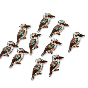 May include: A collection of ten embroidered patches, each depicting a kookaburra bird. The birds feature brown and white plumage with distinct bright blue wing feathers, a large dark beak, and a cream-coloured head, all rendered in detailed thread.
