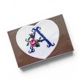 May include: A heart-shaped, white embroidered patch featuring a blue letter "A" and a pink rose design. The patch is on a brown card, suggesting a handmade, personalised item. The stitching is detailed and the colours are vibrant.