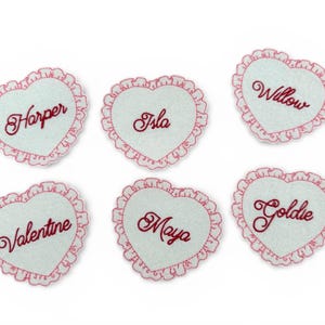 May include: Six heart-shaped patches with names embroidered in red cursive. Each patch has a light blue centre and a pink, ruffled border. Names include Harper, Isla, Willow, Valentine, Maya, and Goldie.