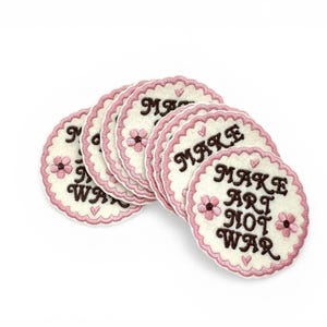 May include: A group of circular patches with a white base and pink, scalloped borders. The patches display the text "MAKE ART NOT WAR" in brown, with pink flower and heart motifs. The patches are arranged on a white backdrop.