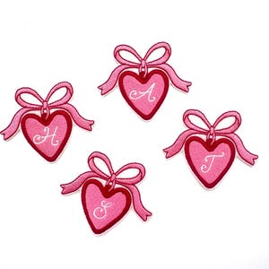 May include: Four pink felt heart-shaped patches with white ribbon bows and white embroidered letters A, H, T, and S.