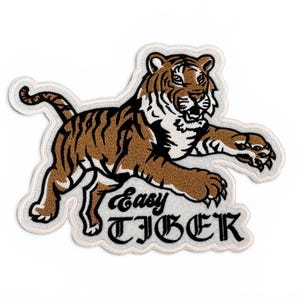 May include: Embroidered patch depicting a brown and black tiger in a running pose. The patch has a white border and the words "Easy TIGER" in black script. The tiger's stripes and details are clearly defined.