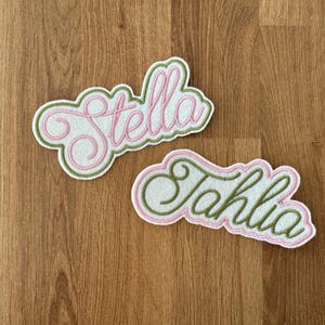 May include: Two white felt patches with pink and green embroidered lettering. The top patch says "Stella" and the bottom patch says "Tahlia".