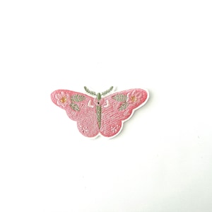 May include: Pink embroidered butterfly with green floral details. The butterfly has a delicate, feminine design.