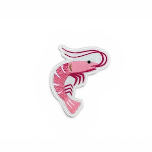 May include: An embroidered patch in the shape of a shrimp. The shrimp is depicted in shades of pink and white with a small black eye and dark pink striped detailing along its curved body. The patch features a white border and a textured finish.