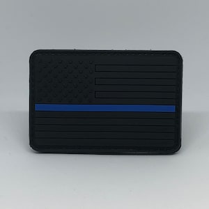 以下が含まれることがあります： 黒と青のアメリカ国旗のパッチで、中央に細い青い線が走っています。パッチは質感のある素材で作られており、ステッチされた縁取りがあります。