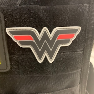 Op de afbeelding: Een grijs en rode Wonder Woman logo-patch met een witte rand en stiksels. De patch is bevestigd aan een zwarte tactische vest.