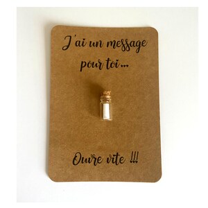 May include: A small clear glass bottle with a cork stopper containing a rolled white paper note, mounted on a rectangular brown card with the black printed text J'ai un message pour toi... and Ouvre vite !!! centered above and below the bottle.