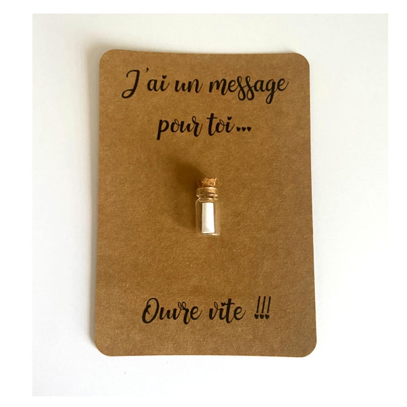 May include: A small clear glass bottle with a cork stopper containing a rolled white paper note, mounted on a rectangular brown card with the black printed text J'ai un message pour toi... and Ouvre vite !!! centered above and below the bottle.