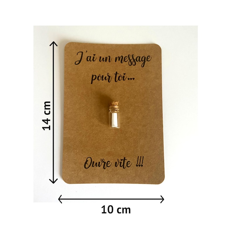 May include: A small glass bottle with a cork stopper containing a white paper scroll, attached to a rectangular brown card measuring 14 cm by 10 cm. The card features the black printed text J'ai un message pour toi... and Ouvre vite !!!
