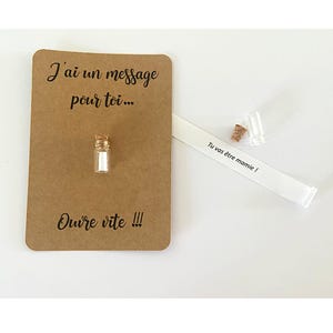 May include: A rectangular brown card with the black text J'ai un message pour toi... and Ouvre vite !!!. A small clear glass bottle with a cork stopper is attached to the card. A white paper strip with the text Tu vas &ecirc;tre mamie ! is displayed.