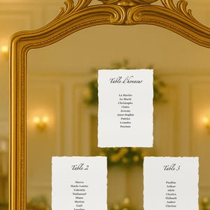 Peut inclure: Un miroir doré encadre trois cartons blancs avec des noms calligraphiés sous les titres "Table d'honneur", "Table 2" et "Table 3". L'ensemble évoque une décoration de mariage ou d'événement, avec une lumière douce.