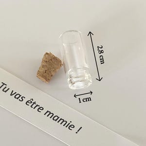 May include: A small clear glass vial with a separate cork stopper. The vial measures 2.8 cm in height and 1 cm in width. A white paper strip is visible with the black printed text: Tu vas &ecirc;tre mamie ! The object is presented for clear size reference.
