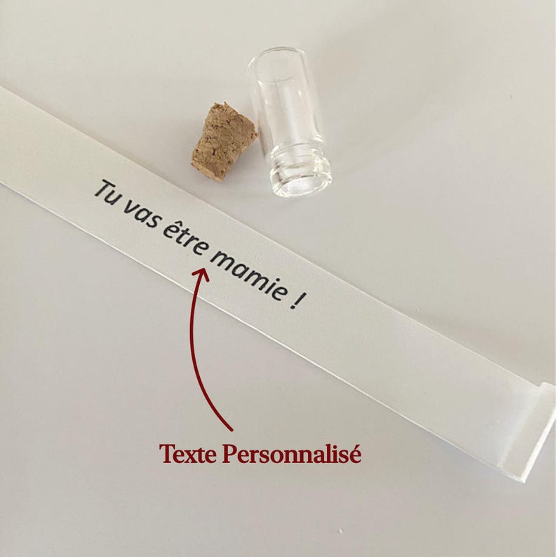 May include: A small clear glass vial with a separate cork stopper and a long white paper strip featuring the black printed text Tu vas &ecirc;tre mamie ! and a red arrow pointing to the text with the red label Texte Personnalis&eacute; below it.