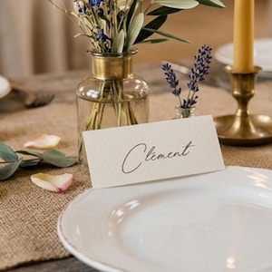 Peut inclure: Un marque-place rectangulaire blanc cassé portant le prénom Clement écrit en écriture cursive noire. La carte est placée devant un vase en verre transparent contenant des fleurs séchées et un bougeoir en laiton avec une bougie jaune.