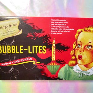 May include: A rectangular box for Bubble-Lites featuring a vintage illustration of a blonde person in a green dress. The box displays the text Nostalgia Series, Bubble-Lites, and Watch Them Bubble, along with technical specifications in inches.