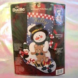 May include: A Bucilla felt stocking kit featuring a snowman and teddy bear design. The packaging displays a blue stocking with a snowman wearing a black top hat, a brown teddy bear, a small penguin, and a candy cane. The text reads Snowman & Teddy Bear.