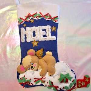 May include: A blue felt Christmas stocking featuring the white stitched text NOEL, a sleeping tan bear with gold wings resting on a white cloud, and decorative red ribbons, green holly leaves, and gold star sequins throughout the design.