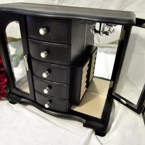 May include: Black jewelry armoire with a mirrored door and four drawers with pearl-like knobs. The armoire has a hinged door on the right side and a small cabinet with multiple compartments. A bouquet of red flowers is visible on the left.