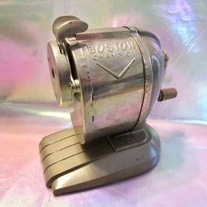 May include: A vintage silver-toned Boston Champion pencil sharpener. The sharpener has a crank handle and a base with a series of horizontal ridges. The words "BOSTON CHAMPION" are visible on the front. A classic office supply.