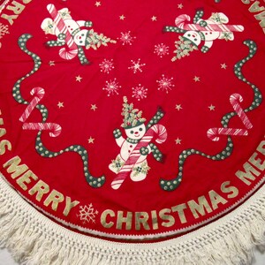 May include: A round red Christmas tree skirt with a white fringe. The skirt features gold lettering that says "MERRY CHRISTMAS" and has illustrations of snowmen, candy canes, and snowflakes.