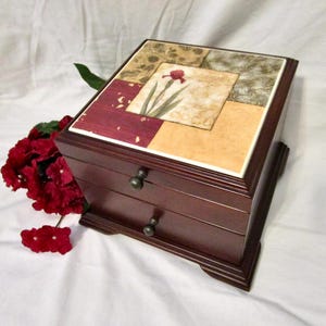 May include: A dark brown wooden jewelry box with two drawers and a decorative top. The top features a floral design with red, beige, and green squares. The box has small, round drawer pulls.