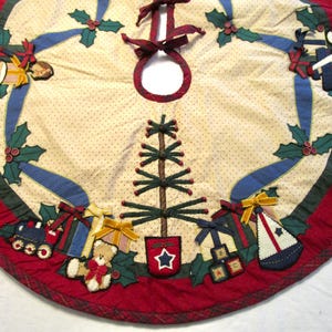 May include: Circular Christmas tree skirt with a cream background and tan spots. It has a red border, green holly leaves, blue ribbons, and appliqued toy and gift designs. A tree is in the centre.
