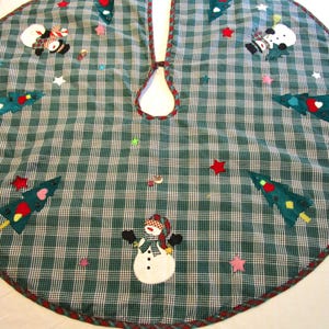 May include: A circular Christmas tree skirt featuring a dark green and white plaid pattern. The fabric is decorated with appliqued snowmen wearing scarves, green Christmas trees with colorful shapes, and scattered red and pink fabric stars.