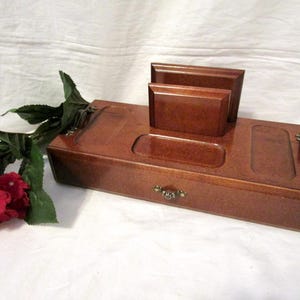 May include: A wooden desk organizer with a drawer, two rectangular compartments, and a small card holder. The organizer is a warm brown color with brass-colored hardware. Red flowers and green leaves are in the background.
