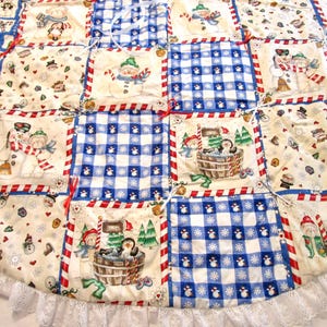 May include: A Christmas tree skirt with a patchwork design featuring snowmen, penguins, and snowflakes. The squares alternate between a cream background with holiday motifs and blue and white gingham. A white lace trim edges the skirt.