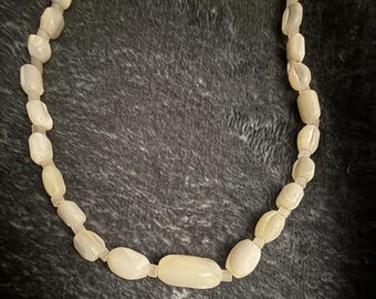 May include: A necklace composed of smooth, off-white, cylindrical beads with a pearlescent finish. The beads are strung together in a single strand, featuring a slightly larger, elongated bead positioned at the center of the necklace design.