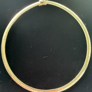 May include: A gold-toned omega chain necklace featuring a smooth, semi-rigid circular design with a polished finish. The necklace is constructed from interconnected flat metal segments and includes a secure, integrated clasp mechanism at the top.