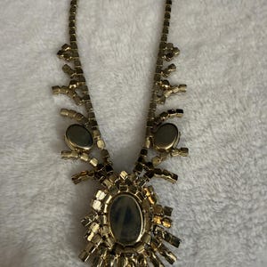 May include: A gold-toned necklace featuring a central oval-shaped pendant surrounded by radiating rectangular metal segments. The chain consists of a series of connected square links, with two additional oval accents positioned along the sides.