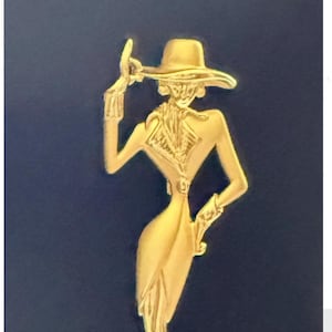 Könnte beinhalten: Eine goldfarbene Metallbrosche in Form einer stilisierten Figur mit breitkrempigem Hut und Schal. Die Figur hat einen Arm zum Hutrand erhoben und den anderen in die Taille gestützt, mit glatten, länglichen Linien und einer polierten Oberfläche.