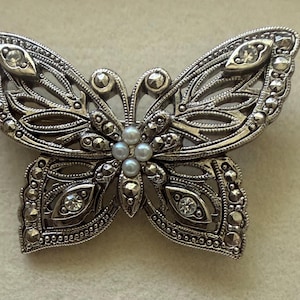 May include: A silver-toned butterfly brooch featuring intricate openwork metal detailing, accented with small round white pearls at the centre, sparkling clear crystals on the wings, and numerous dark grey marcasite stones throughout the frame.