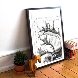 May include: Black framed art print with a detailed pen and ink illustration of a monster with antlers and text. The artwork is propped against a white wall on a wooden floor, with a small potted plant and books nearby.